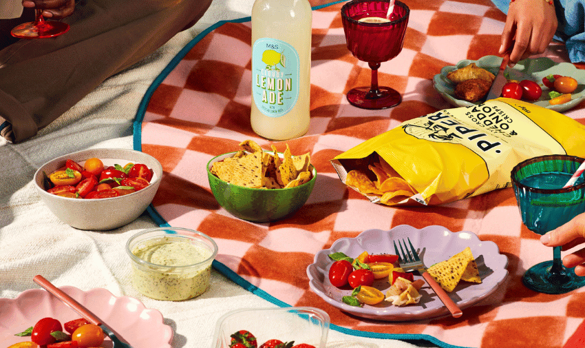 A colourful picnic spread on a round blanket features a variety of food and drinks, including a bottle of lemonade, dishes of salad and veggies, a bowl of tortilla chips, and vibrant cups. Three people seated around the blanket are visible.