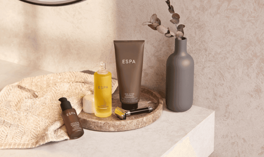 A minimalist bathroom scene featuring ESPA-branded skincare products on a round stone tray. The products include a brown tube, a yellow liquid in a glass pump bottle, and a smaller brown bottle. A beige towel and a tall, matte grey vase with eucalyptus branches are nearby.