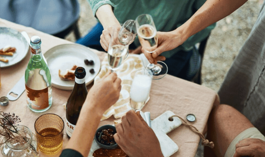 Five people clinking their glasses of champagne over a table set with drinks, plates, and snacks. Bottles of water and wine, along with an assortment of small dishes, are also on the table. The scene suggests a celebratory or social gathering.