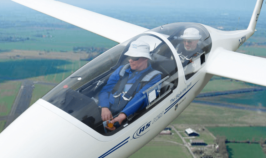 Two people wearing helmets and safety harnesses flying in a two-seater glider, with a panoramic view of green fields and an airstrip visible below. The pilot is in the front seat, operating the controls, while the co-pilot sits behind.