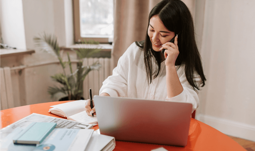 A woman seated at a desk with a laptop, smiling as she speaks on the phone and takes notes, symbolising managing rent payments and building credit effortlessly.