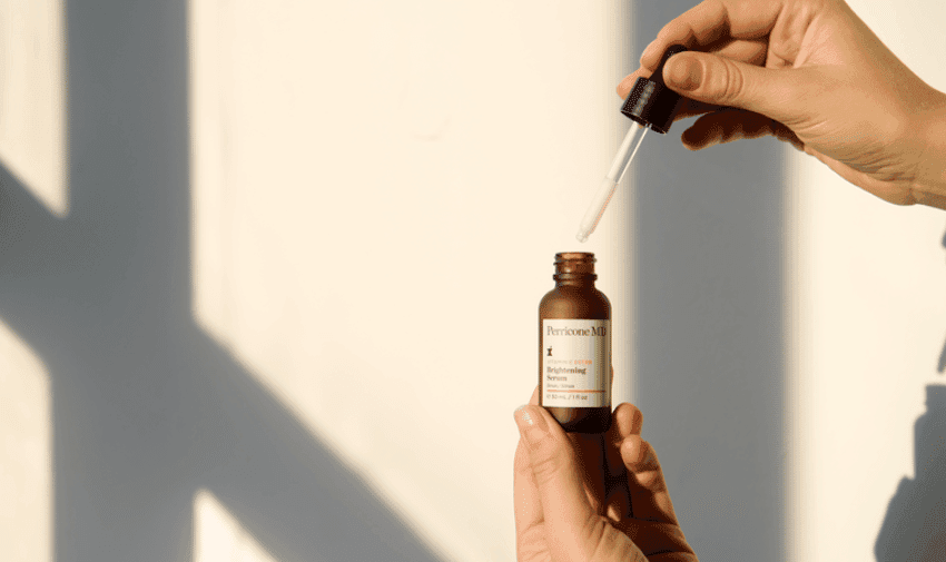 A person holds a small dark brown serum bottle labeled "Primrose Milk" with one hand and an eyedropper with a drop of liquid in the other hand. The background is out of focus with soft natural light casting geometric shadows on a light-coloured wall.