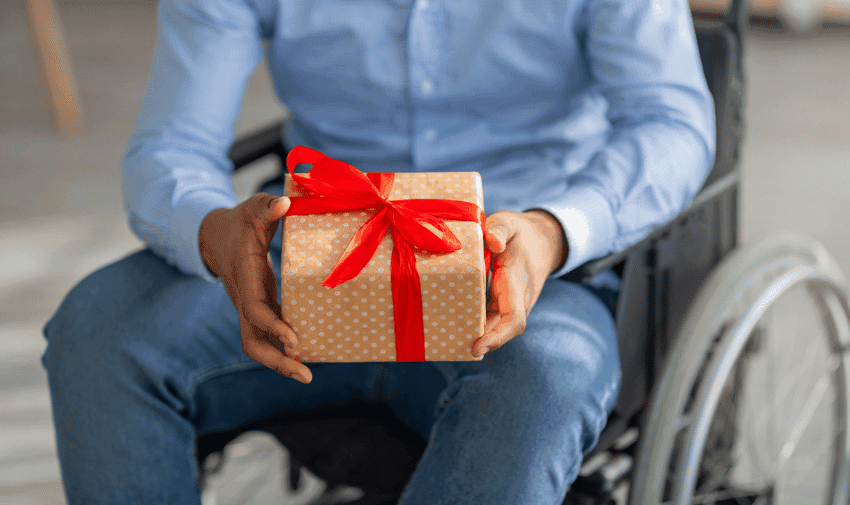 A person in a wheelchair holds a wrapped gift with a red ribbon and bow in their hands. The person wears blue jeans and a light blue shirt. The background is blurred, focusing on the gift and the person's hands.