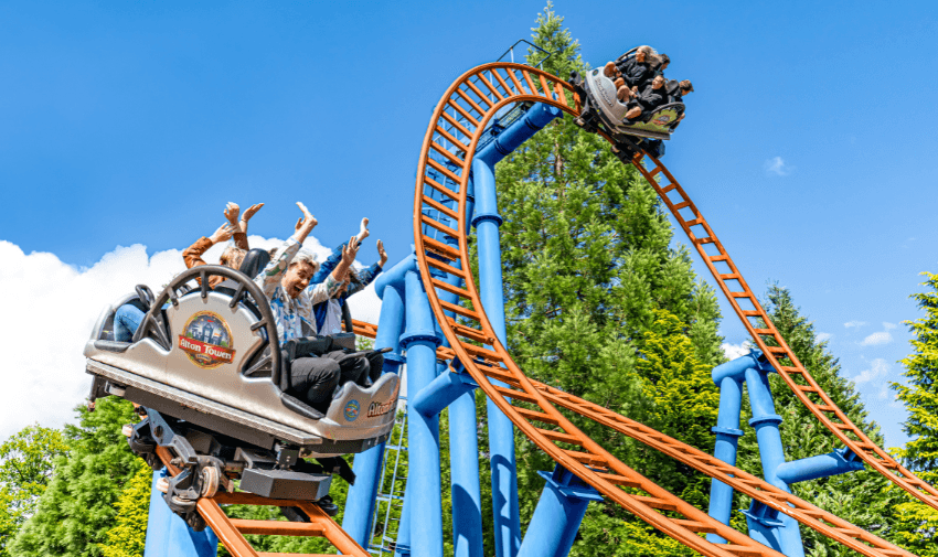 Thrill-seekers riding a roller coaster with vibrant orange tracks and blue supports, hands raised in excitement against a clear blue sky.