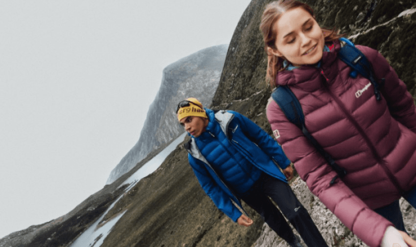 Two people hiking on a mountain trail, dressed in warm jackets and backpacks. The woman in a maroon jacket leads, followed by someone in a blue jacket and yellow headband. The landscape is rugged with overcast skies.