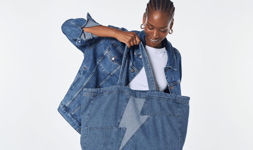 A person wearing a blue denim jacket with their hair in braids is smiling and looking down while holding a large, oversized denim bag. The background is plain white.