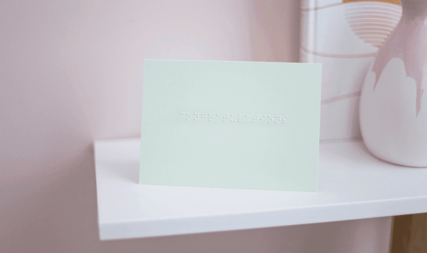 A pastel green card with congratulations written in lower case braille. The card is on a white shelf with a vase and print just visible on the right.