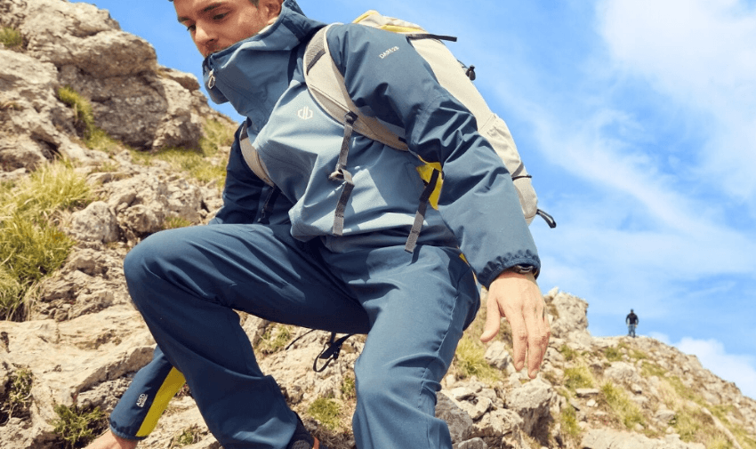 A person wearing a blue hiking outfit and backpack climbs a rocky hill under a bright blue sky. In the background, another individual stands atop the rocks, enjoying the scenic view.