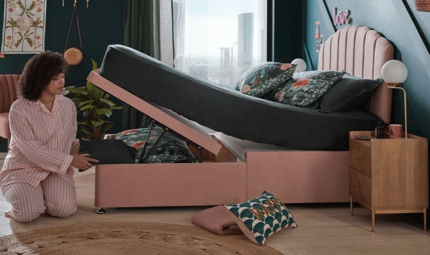 A stylish bedroom featuring a pink ottoman bed with under-bed storage lifted open. A woman in pink striped pyjamas kneels beside it, placing items inside. The room is decorated with plants and modern furniture. Shopping Discounts for Disabled People, Accessible Home Discounts UK.