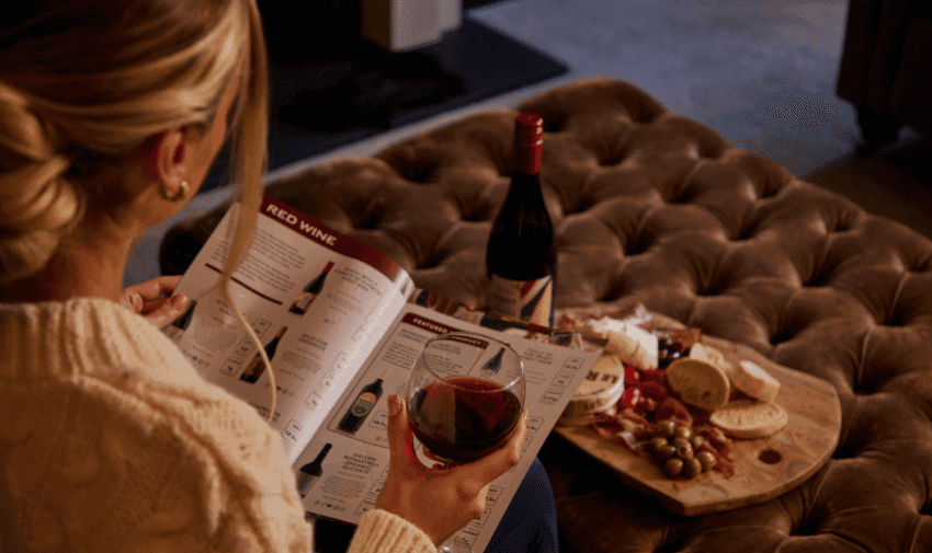 A woman with blonde hair reads a wine catalogue while holding a glass of red wine in her left hand. A bottle of red wine and a wooden platter with assorted cheese, olives, and charcuterie are on a brown ottoman in front of her.