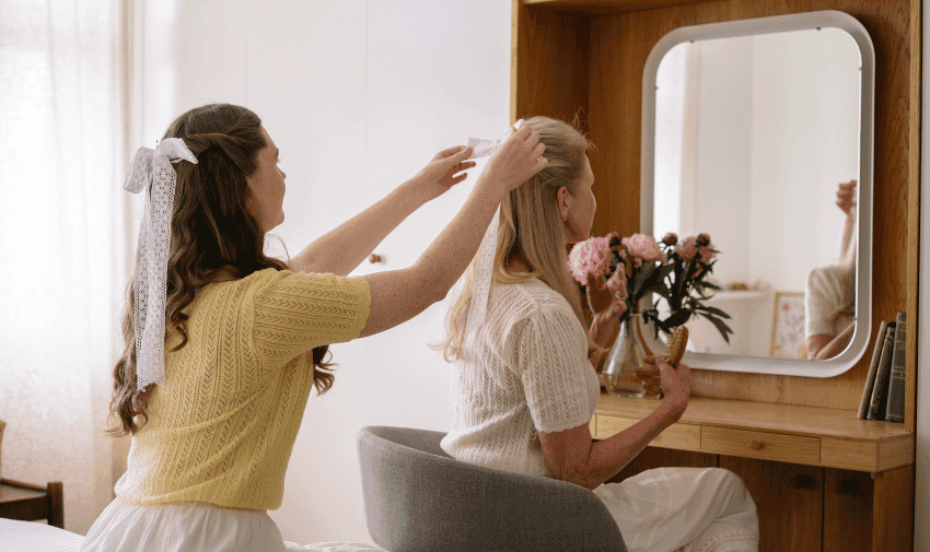  A young woman with a white bow in her hair is placing a hair accessory on an older woman with long blonde hair who is seated at a dresser with a mirror. The dresser holds a bouquet of flowers and some books. The room appears cozy and well-lit.