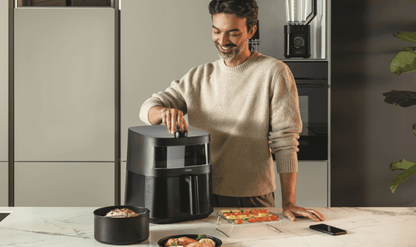 A smiling individual is using a black air fryer on a kitchen counter. Nearby, there are prepared food items, including a bowl of ingredients and a plate of cut vegetables. The kitchen has modern appliances and a tidy look