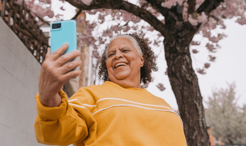 An elderly woman in a yellow sweatshirt smiles joyfully while taking a selfie with her smartphone. She's standing under a blooming cherry blossom tree, enjoying a bright, cheerful day.