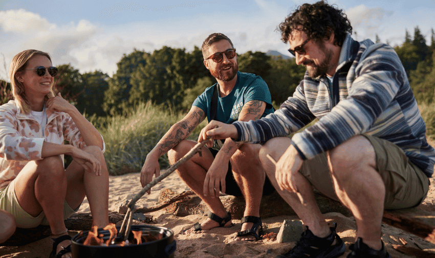 Three people, wearing casual summer attire and sunglasses, are sitting on a sandy beach around a small campfire. One person is tending to the fire with a stick. They are laughing and enjoying the sunny day, with trees and clear skies in the background.