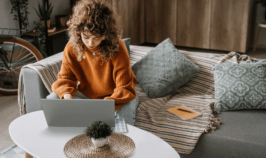 A person with curly hair sits on a couch, focused on their laptop placed on a white coffee table. Nearby, there is a notebook and a potted plant.