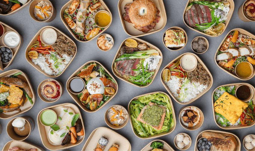 An array of neatly plated meals in eco-friendly containers, featuring diverse dishes like grilled meats, fresh vegetables, sushi, and desserts, arranged on a grey surface.