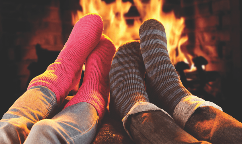 Two pairs of feet in cosy socks, one red and one striped, rest on a surface in front of a warm, glowing fireplace, creating a cozy and relaxing atmosphere.