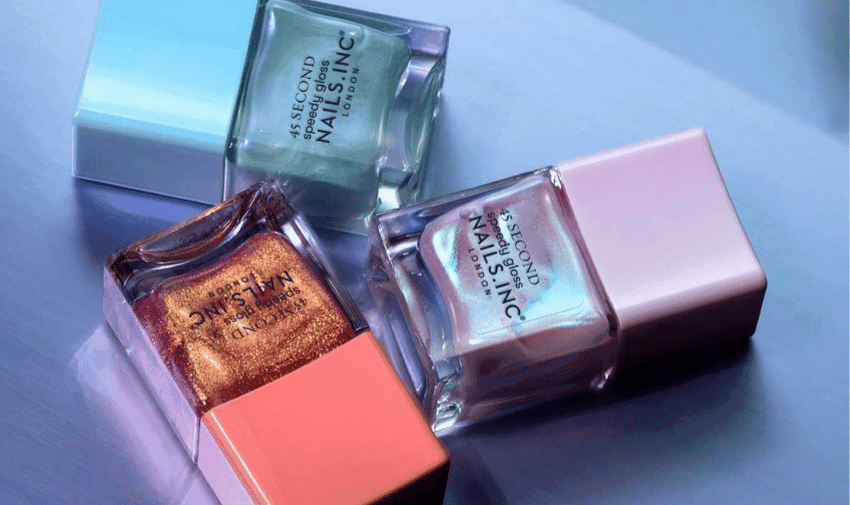 Three bottles of Nails Inc nail polish are arranged on a reflective surface. The bottles are laid out in a triangular formation, showcasing colours including metallic gold, iridescent purple, and aqua with a shiny finish. All bottles have pale, pastel-coloured caps.