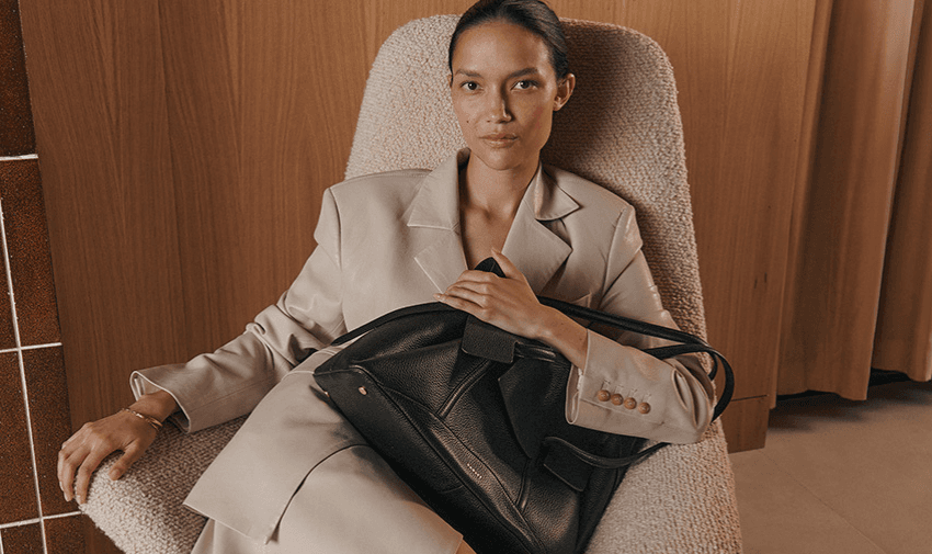 A person in a beige suit sits in a textured chair, holding a large black handbag. The setting includes wooden panels and a tiled wall, creating a modern and elegant atmosphere.