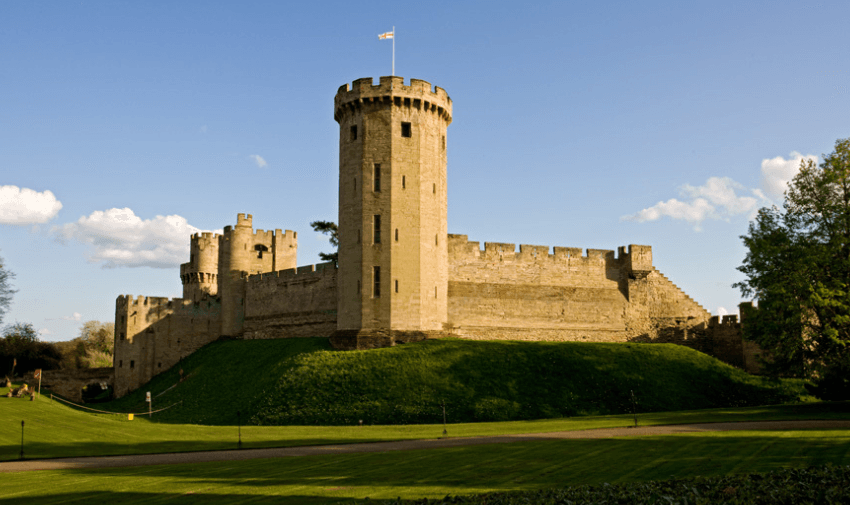 Warwick Castle, a historic stone fortress with a tall tower flying the English flag, set against a bright blue sky and lush green grounds. Highlights Heritage Discounts for Disabled People and Accessible Travel Offers.