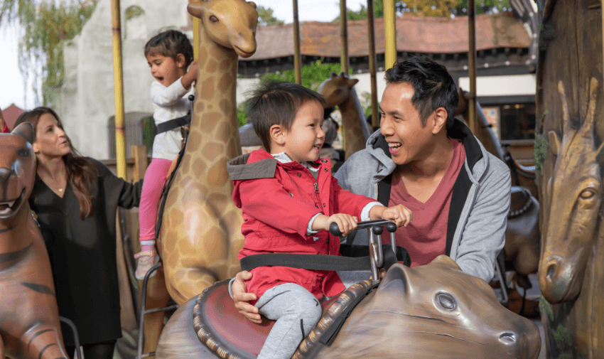 A father and young child smiling together on a carousel ride with animal-shaped seats, surrounded by other families enjoying the ride.