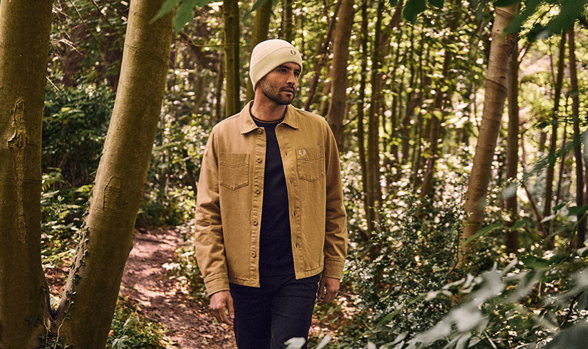 A person wearing a beige jacket and white beanie walks through a forested area, surrounded by tall trees and greenery, creating a peaceful natural setting.