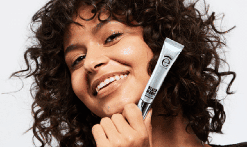 A person with curly hair smiles broadly while holding Eyeko "Black Snail" near their face. The background is plain white.