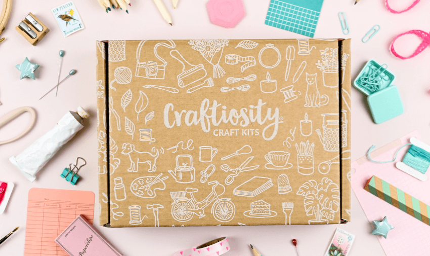 Craftiosity craft kit box surrounded by colourful craft tools and supplies on a pastel pink surface. highlights hobby discounts for disabled people and craft gift offers.