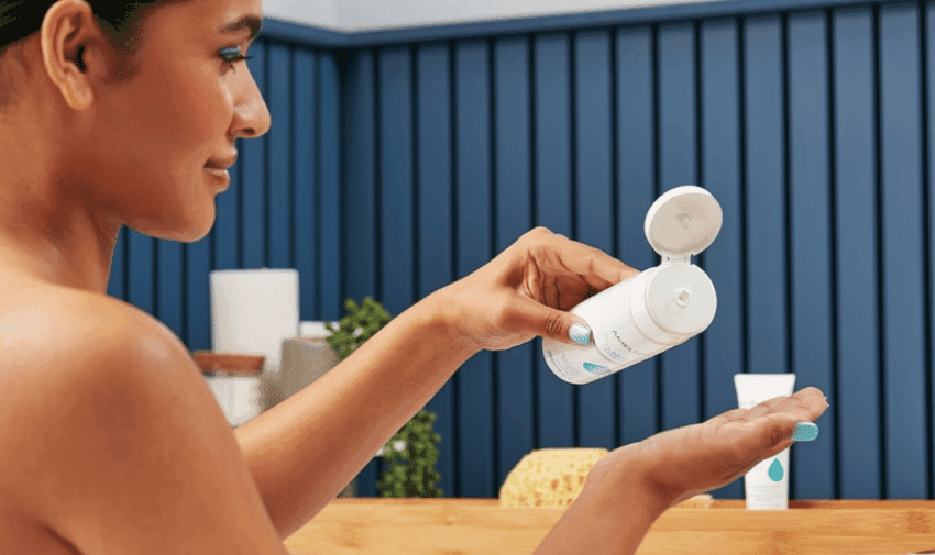 A person with a towel wrapped around their chest is seen dispensing lotion from a white bottle onto their palm. They are in a bathroom with blue paneling, a wooden counter, and other skincare products in the background.