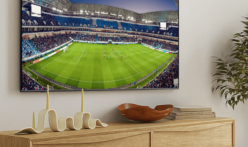 A wall-mounted TV displaying a live soccer match in a packed stadium. Below the TV is a modern white cabinet with decorative items, including a unique candle holder, a wooden bowl, and several stacked books. A potted plant stands nearby.