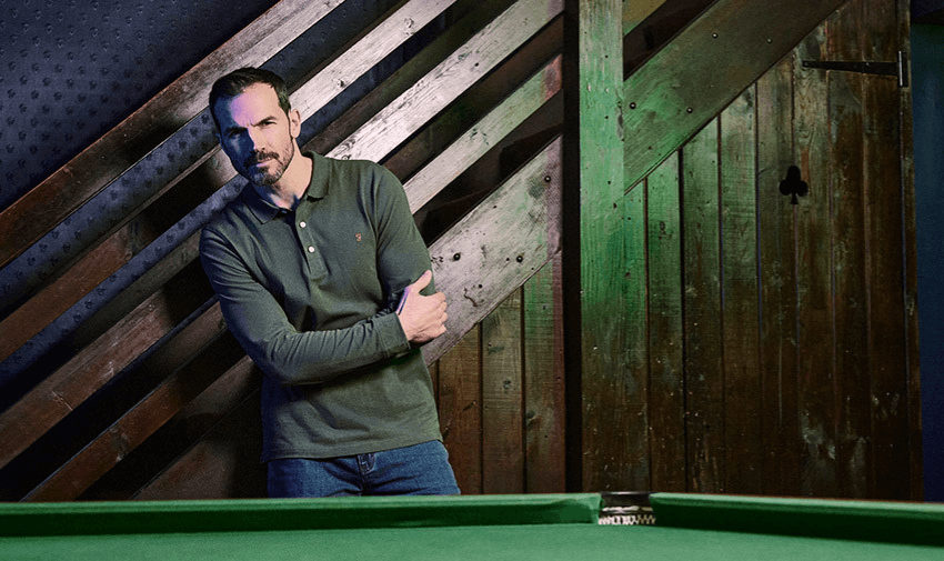 A man wearing a stylish green polo shirt, leaning against a rustic wooden backdrop, showcasing Mainline Menswear. Highlighting Purpl's exclusive disabled discounts for premium men's fashion in the UK.