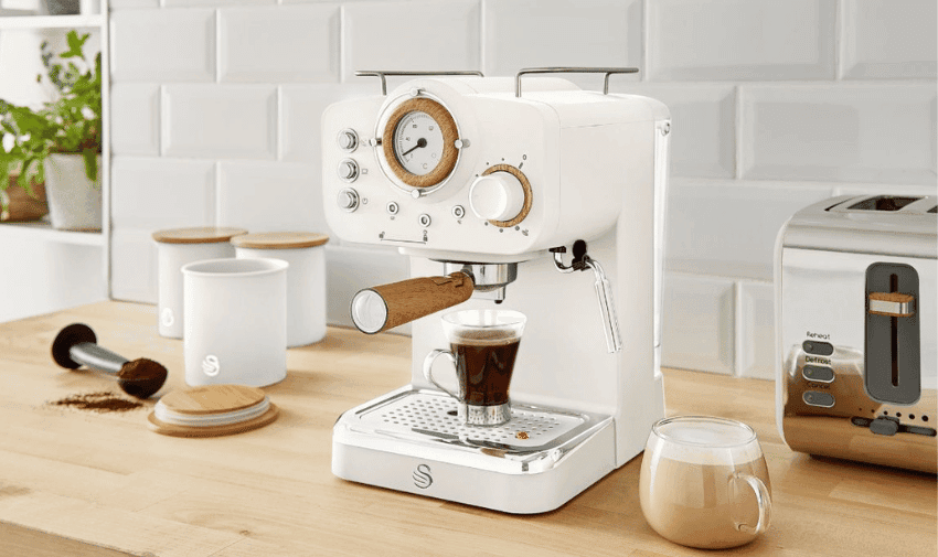 A modern white SWAN espresso machine with wooden accents sits on a kitchen counter. It dispenses coffee into a glass cup. Nearby are two white canisters with wooden lids, another cup of coffee with milk, and a stainless steel toaster. A potted plant is in the background.