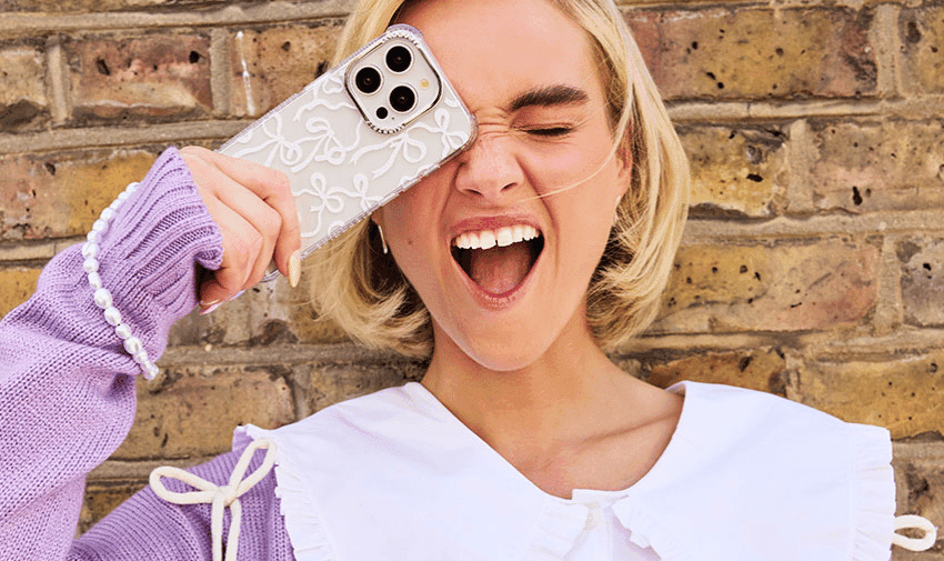 A woman with blonde hair, smiles widely with one eye closed while holding a smartphone with an intricate white lace-like design case against their face. She is standing in front of a brick wall.