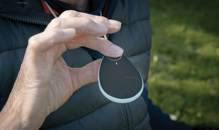 A close-up of an elderly person holding a black and white teardrop-shaped personal alarm device, worn on a lanyard. The person is dressed in an outdoor jacket, and the background shows a blurred green lawn, suggesting an outdoor setting. The device is designed for safety and emergency assistance