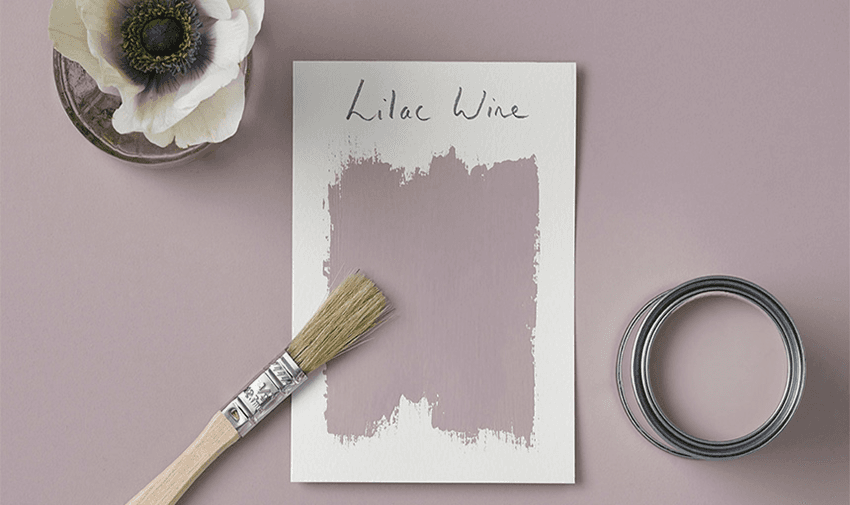 a paint brush laid next to a piece of white paper with a sample of Lilac Wine paint, there is a pot of opened paint next to it and the background is the same colour.