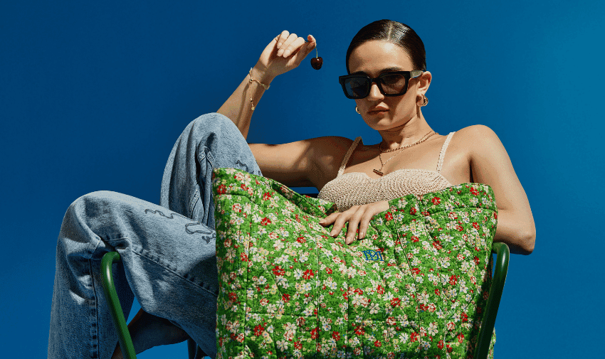 A woman wearing sunglasses, a beige top, and blue jeans sits on a green chair against a blue background. They hold a cherry in one hand and have a large green floral bag on their lap.