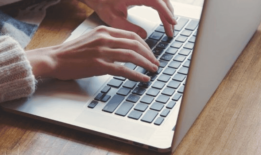 Close-up of a person's hands typing on a silver laptop with black keys. The person is wearing a light-coloured, knitted sweater and is seated at a wooden surface. The lighting is soft, creating a comfortable and relaxed atmosphere.