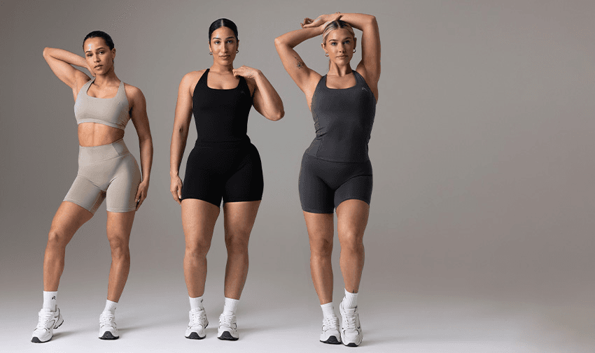 Three women pose confidently in athletic wear against a neutral background. They wear matching Oner Active tops and shorts in beige, black, and dark grey, respectively, with white sneakers. Their varying body types and casual, strong poses promote body positivity and fitness.