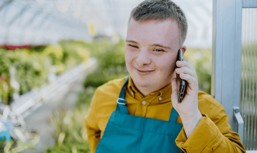 A young man with Down syndrome is smiling while talking on a smartphone. He is wearing a mustard-colored shirt and a teal apron, standing in a greenhouse with blurred plants and gardening supplies in the background.