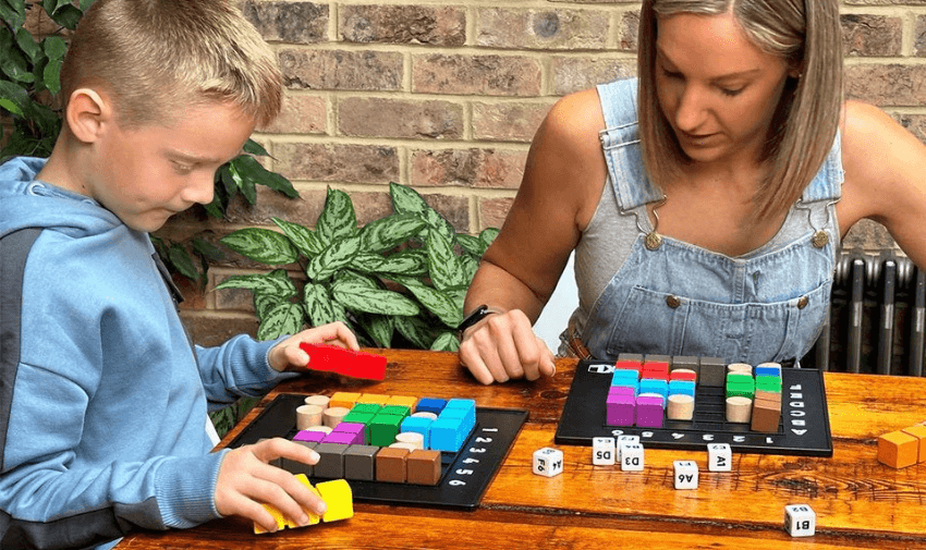 A woman and a boy are sitting at a wooden table playing a board game involving colourful blocks and dice. The boy is holding a yellow block, while the woman focuses on the pieces. A brick wall and a green plant are in the background