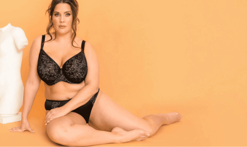 Brastop D-K cup lingerie. The Fuller Bust Outlet. A woman sitting on the floor against an orange background, wearing a black bra and matching briefs with lace details from the brand Adella. A white mannequin torso is visible on her left.