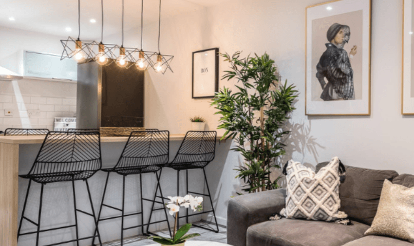 A modern living room and kitchen area with a grey couch adorned with patterned pillows, black wire barstools at a wooden counter, geometric pendant lighting, potted plants, and framed artwork on the walls. An orchid plant sits on the floor near the couch.