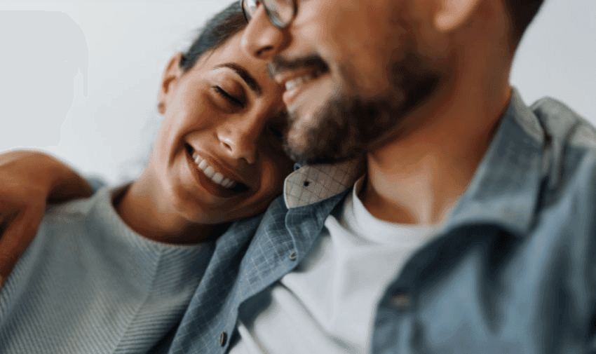 A close-up of a couple embracing. The woman has her eyes closed and smiles contentedly while resting her head on the man's shoulder. The man, wearing glasses and a beard, is also smiling. Both appear relaxed and happy.