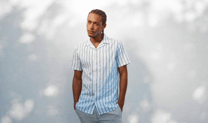 A person with long braided hair stands against a blurred background, wearing a light blue and white vertical striped short-sleeve button-up shirt and light gray pants. The person has their hands in their pockets and is gazing slightly to the side with a relaxed expression.