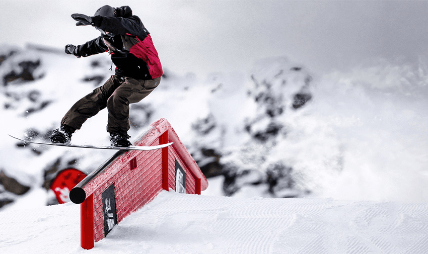 A snowboarder in mid-air performs a trick on a rail in a snowy mountainous terrain. The snowboarder is wearing a red and black jacket, brown pants, and a helmet, with snow-capped peaks and a grey sky in the background.