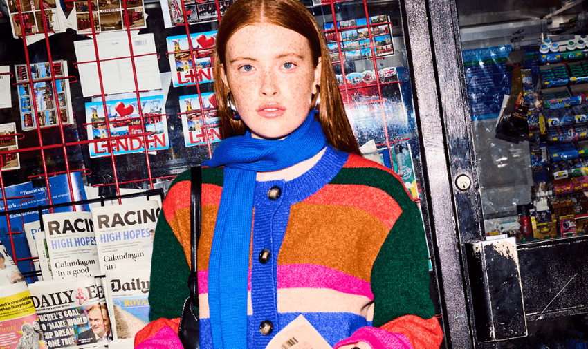 Young woman with striking red hair wearing a colourful striped jumper and vibrant blue scarf, standing outside a London newsagent showcasing modern, sustainable fashion by Omnes