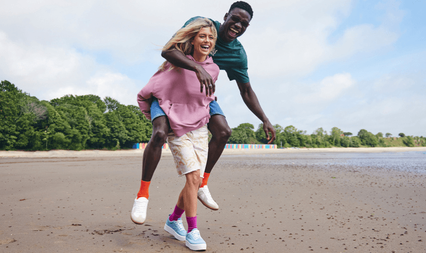 A joyful scene on a sandy beach with a woman in a pink hoodie and a man in a green shirt. She carries him on her back as they both laugh, with colourful beach huts and green trees in the background under a partly cloudy sky.