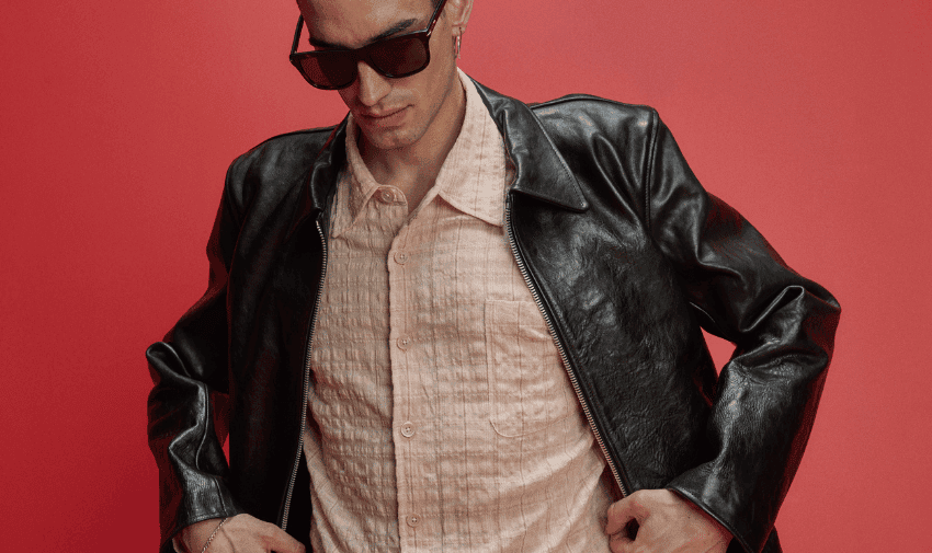 A person wearing a black leather jacket with a light coloured, textured button-up shirt and dark sunglasses stands against a red background.