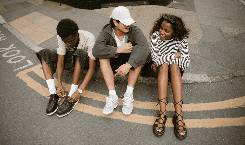 Three people sitting on a double yellow line at the side of a road. One person is tying their shoe, another wearing a cap is sitting in the middle, and the third person with curly hair sits on the right, smiling and hugging their knees.