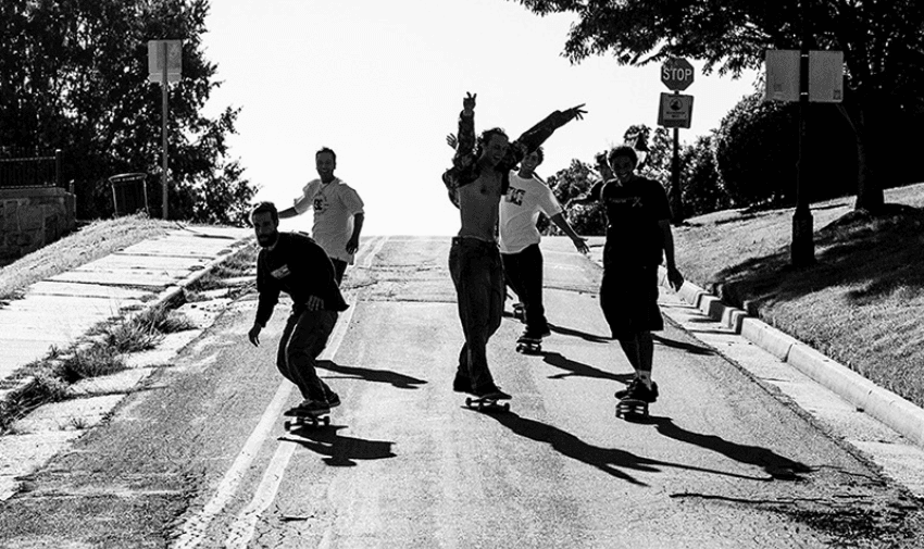 A group of men on skateboards are coming along the centre of a road. It is a black and white image.